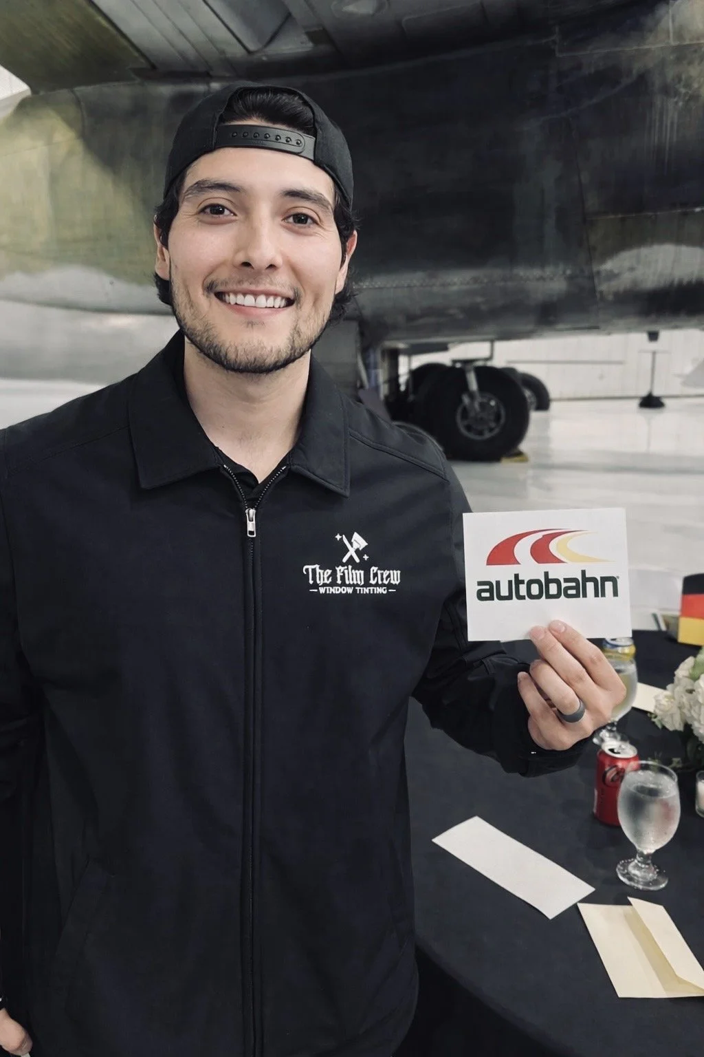 Adrian, founder of The Film Crew Automotive, smiling in a Film Crew jacket and holding an Autobahn dealer card